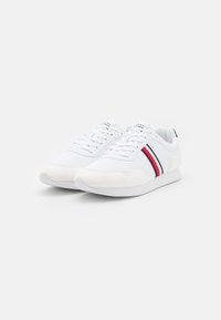 Białe sneakersy z teksturowaną cholewką, płaskimi sznurówkami i gumową podeszwą. Ciekawe detale to czerwone i granatowe paski po boku.