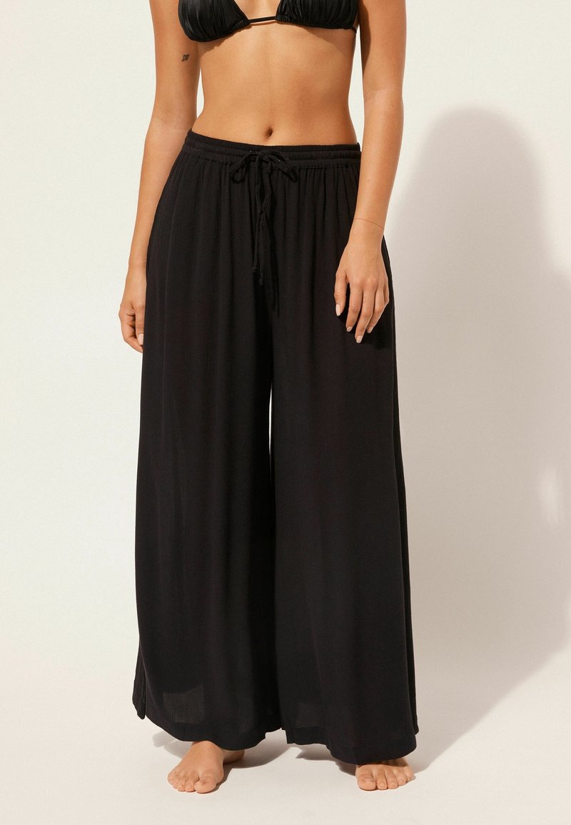 Femme portant un pantalon noir ample à cordon et un haut de bikini noir, debout pieds nus sur un fond uni.