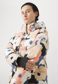 Vadderad jacka med ett flerfärgat abstrakt mönster i rosa, blått och beige. Inkluderar en hög krage och svarta handledsmuddar.