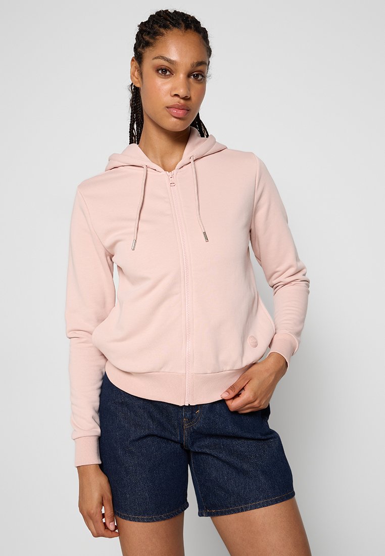 Felpa rosa chiaro con zip in tessuto morbido, caratterizzata da un cappuccio con cordino e polsini a costine. Abbinata a pantaloni denim per un contrasto.