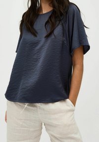 Top blu navy a maniche corte in tessuto leggero e testurizzato, con vestibilità ampia e scollatura arrotondata, abbinato a pantaloni di lino chiari.