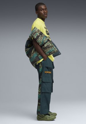Jeune homme debout de profil portant un t-shirt jaune à motif, une veste camouflage verte drapée sur l'épaule, un pantalon cargo et des baskets vertes sur fond gris.