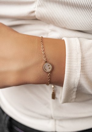 Polso con braccialetto a catena dorata con ciondolo rotondo a forma di faccina sorridente, indossato sopra una maglia bianca a coste a manica lunga.