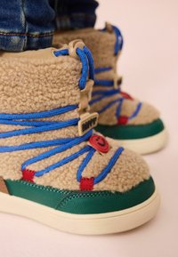 Botte en polaire beige avec lacets bleus, accents rouges, base verte et une étiquette ronde avec un smiley. Surface texturée avec un col rembourré.