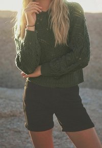 Pull en maille vert avec un motif texturé, col rond et poignets côtelés, associé à un short noir moulant. Éclairage naturel et chaleureux.