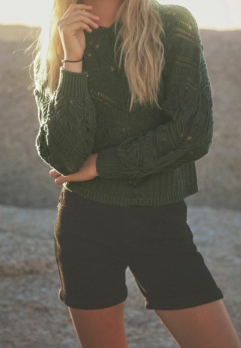 Pull en maille vert avec un motif texturé, col rond et poignets côtelés, associé à un short noir moulant. Éclairage naturel et chaleureux.