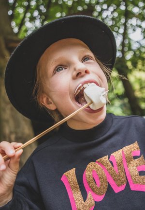 Kind dat een zwarte hoed en een zwart sweatshirt met roze en gouden "LOVE"-tekst draagt, terwijl hij een prikker met een geroosterde marshmallow vasthoudt.