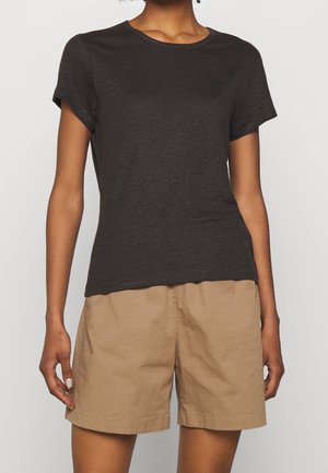 Person trägt ein schlichtes dunkelgraues Kurzarm-T-Shirt und locker sitzende hellbraune Shorts vor einfarbigem Hintergrund.