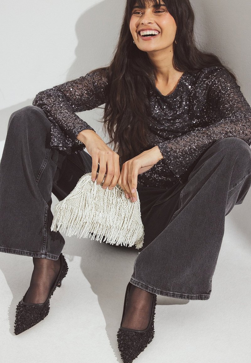 Tacones negros de punta con cuentas, pantalones anchos de denim gris sueltos y un bolso de cuentas de flecos blancos, combinados con una blusa negra brillante.