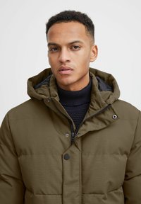 Giacca puffer verde oliva con chiusura con zip, cappuccio e maglione nero a coste sotto. Presenta un tessuto liscio e testurizzato con sezioni cucite.