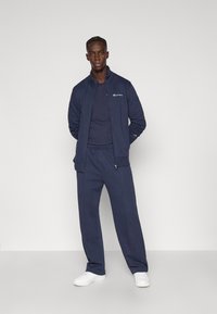 Σκούρο μπλε φόρμα με ζακέτα με φερμουάρ και παντελόνι με ελαστική μέση. Κατασκευασμένη από απαλό ύφασμα, διαθέτει λογότυπα και συνδυάζεται με λευκά αθλητικά παπούτσια.