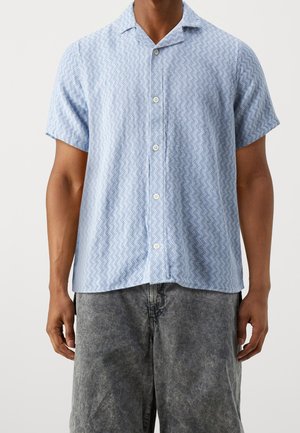 Homme portant une chemise bleu clair à manches courtes avec un motif en zigzag et un short en jean noir délavé.