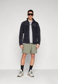 Temno siva flis jakna z zadrgo spredaj, nošena preko svetlo sive majice in zelenih kratkih hlač. Model dopolnjen s črnimi nogavicami in supergami.