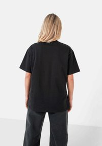 Camiseta de algodón negra con un corte relajado, mangas cortas y escote redondo, vista desde la parte de atrás, combinada con pantalones oscuros de pierna ancha.