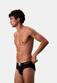 Schwarze Baumwollslip mit weißem Logobund, mit angepasstem Schnitt und glatter Textur, an einem männlichen Modell im Profil dargestellt.