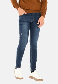 Jeans skinny in denim blu scuro con lievi strappi sulla coscia. Indossati con sneakers bianche e un maglione marrone.