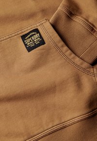Felpa in cotone marrone con colletto a coste testurizzato e un'etichetta con la scritta "Superdry Workwear" nella zona della tasca anteriore.