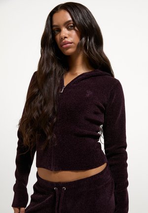 Frau mit langen dunklen Haaren, die einen dunkelvioletten, gekürzten Hoodie mit Reißverschluss und passende Hosen trägt, stehender vor einem einfarbigen hellen Hintergrund.