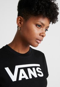 Musta puuvillateepaita, jossa on suuri valkoinen "VANS"-logo. Pyöreä kaula-aukko, lyhyet hihat ja sileä pinta.
