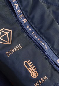 Marineblåt stof med kobberfarvede detaljer, med teksten "DURABLE" og "WARM" samt et ikon af et termometer. Broderet mærkenavn synligt.
