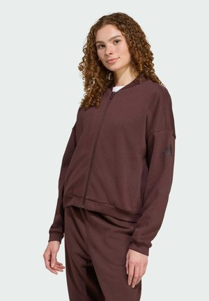 Jeune femme aux cheveux bouclés portant une veste zippée marron et un pantalon assorti, debout devant un fond gris clair uni.