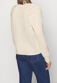 Beige sweatshirt med rynkade axeldetaljer, mjuk textur, ribbade ärmslut och en avslappnad passform, i kombination med mörkblå jeans.