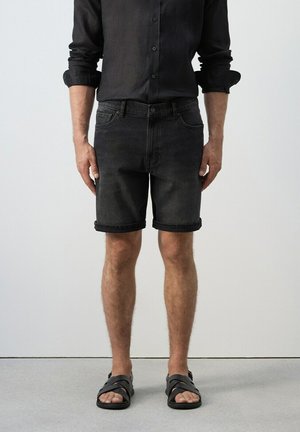 Hombre con camisa negra de botones, pantalones cortos de mezclilla con dobladillo enrollado y sandalias negras de correas cruzadas, de pie sobre un piso gris.