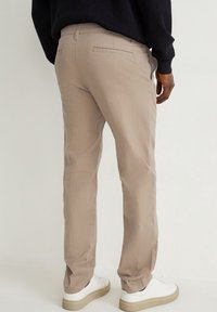 Homme portant un pantalon chino beige, des baskets blanches et un pull noir, debout de dos devant un fond blanc uni.
