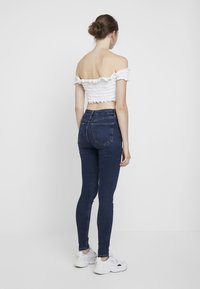 Vit off-the-shoulder smockad crop top med volangkanter, parat med högmidjade mörkblå skinny jeans och vita sneakers.