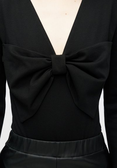 Μαύρο ολόσωμο ρούχο με βαθύ V λαιμό και μεγάλη μαύρη λεπτομέρεια φιόγκος στο στήθος, φτιαγμένο από ομαλό, ελαστικό ύφασμα.