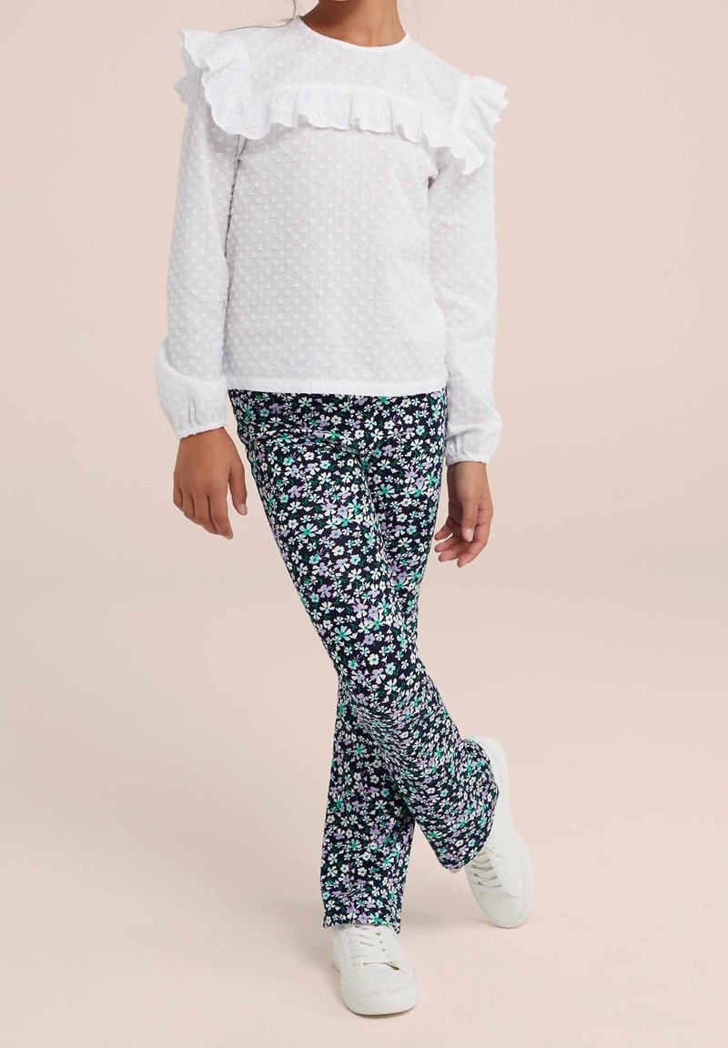 Enfant portant un chemisier blanc texturé à manches longues avec des volants et un pantalon noir à motif floral avec des baskets blanches.