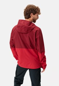 Veste imperméable rouge avec capuche, présentant un design bicolore. Fabriquée en matériau lisse, elle a une coupe légèrement ample avec des poignets élastiques.