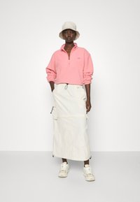Sudadera de felpa rosa claro con cremallera y mangas largas, combinada con una falda larga blanca con bolsillos y cordones, y un sombrero de cubo beige.