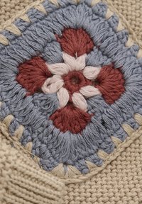 Baumwolltextil mit einem blauen Häkelquadrat und floralen Mustern in Rot und Pink, umrahmt von crèmefarbenem Garn, auf einem strukturierten beigen Hintergrund.
