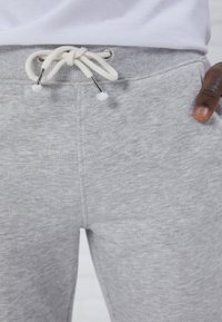 Grå joggebukser har en myk tekstur, ribbestrikket midjebånd og hvit snor med metalløyer. Stoffet har en subtil spraglete effekt.