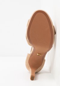 Chaussure à talon haut en daim beige vue de la semelle, présentant un talon fin et un petit logo doré près de la voûte plantaire.