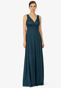 Lungo vestito di teal con corpetto in pizzo, scollatura a V e gonna fluente. Presenta spalline larghe e vita aderente, dettagli testurizzati sulla parte superiore.
