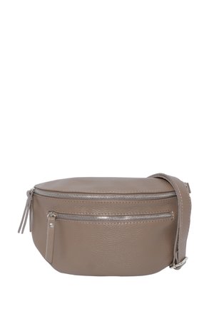 Junge Frau lächelnd, trägt ein schwarzes langärmeliges Oberteil und Jeans, trägt eine taupefarbene Leder-Umhängetasche mit Reißverschlusstaschen.