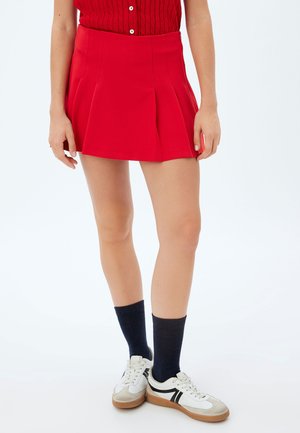 Rode geplooide rok van gladde stof, met een hoge taille. Gecombineerd met witte sneakers en donkere sokken. Achtergrond is vlak wit.