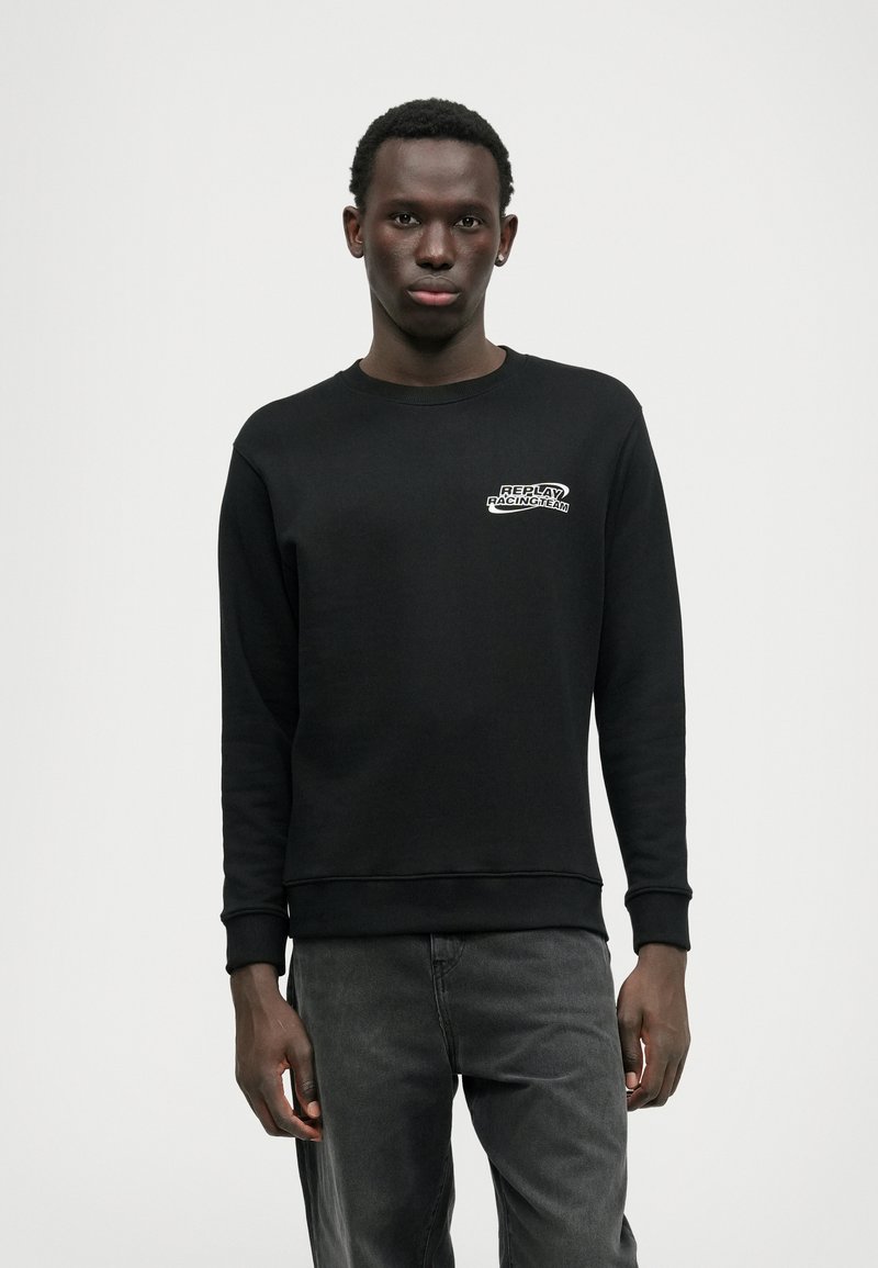 Sweatshirt preto com gola redonda, punhos e bainha canelados. Apresenta um patch com logotipo branco no peito. Tecido macio e texturizado, ajuste descontraído.