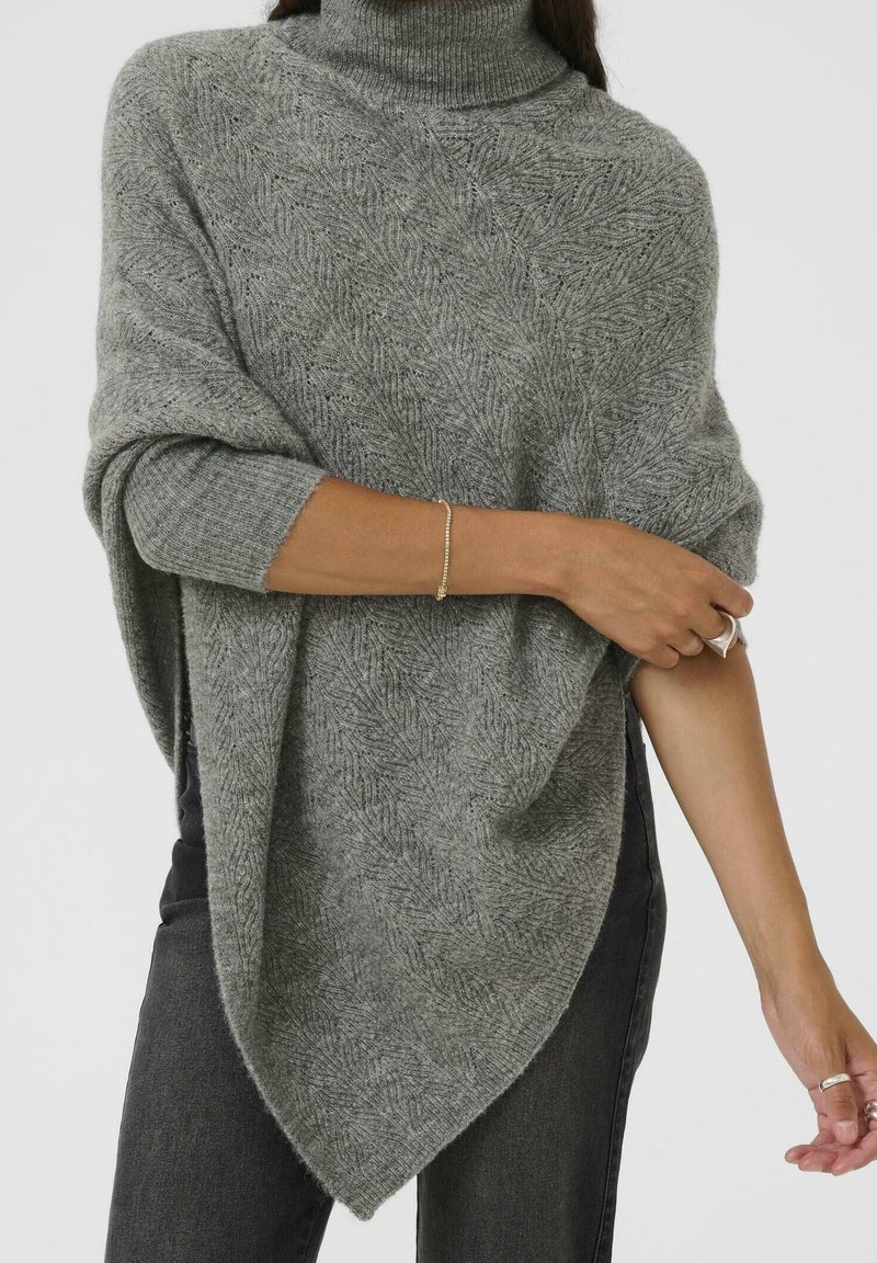 Femme portant un poncho en maille texturée grise avec col roulé et manches côtelées, associé à un jean foncé et des bijoux argentés aux mains et au poignet.