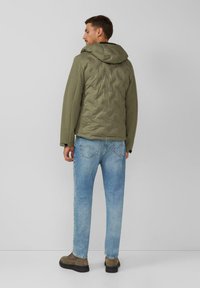 Kurtka pikowana w kolorze oliwkowej zieleni z kapturem, ozdobiona przeszyciami; zestawiona z jasnoniebieskimi dżinsami i brązowymi botkami do kostki.