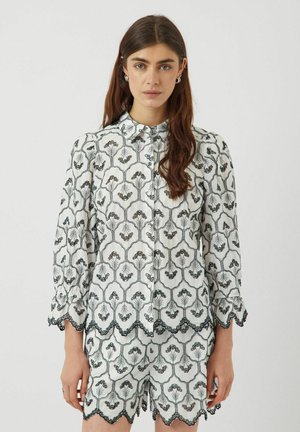 Chemise blanche avec une broderie florale noire complexe, devant boutonné et manches à bords festonnés. Associée à un short assorti présentant le même motif.