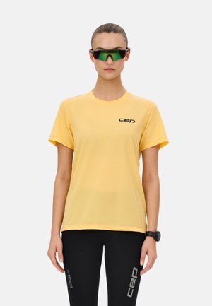 Femme portant un t-shirt CEP jaune, des leggings CEP noirs, des lunettes de sport réfléchissantes vertes et une montre-bracelet noire, se tenant devant un fond blanc.