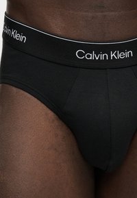 Μαύρα ανδρικά εσώρουχα με λευκό λογότυπο Calvin Klein στη μέση, φορεμένα σε σκούρο δέρμα, δείχνοντας την περιοχή των γοφών και του άνω μηρού.