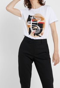 T-shirt en coton blanc présentant un design graphique abstrait en rouge, noir et marron. Associé à un pantalon noir bien ajusté.