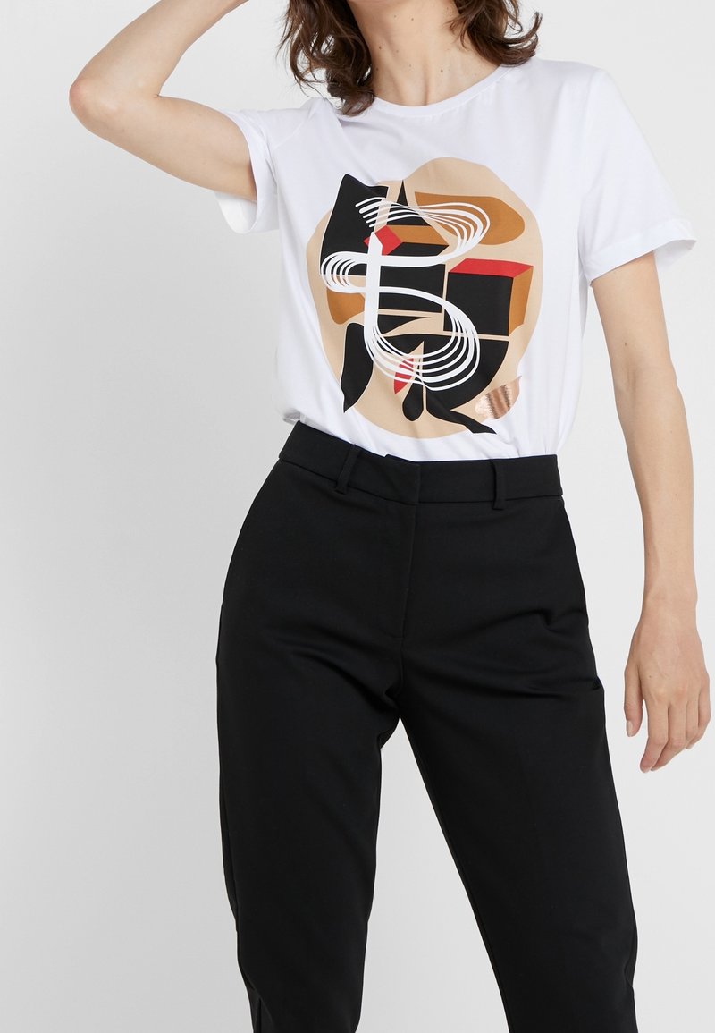 T-shirt en coton blanc présentant un design graphique abstrait en rouge, noir et marron. Associé à un pantalon noir bien ajusté.