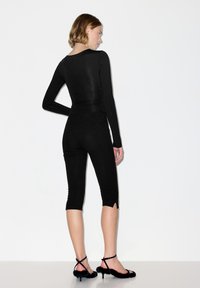 Vrouw in een zwart shirt met lange mouwen en nauwsluitende cropped broek, draagt zwarte fluffy riemhakken, staand tegen een witte achtergrond zijwaarts gericht.