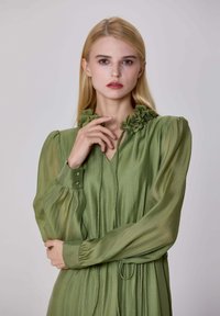Femme blonde avec des cheveux raides, portant une robe verte à manches longues avec des détails floraux sur le col, posant devant un fond uni.