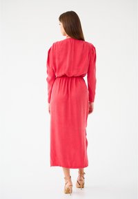 Robe maxi en lin rouge avec une taille cintrée, des manches longues et des fentes sur les côtés. Portée avec des talons beiges à lanières, présentée sur un fond blanc.